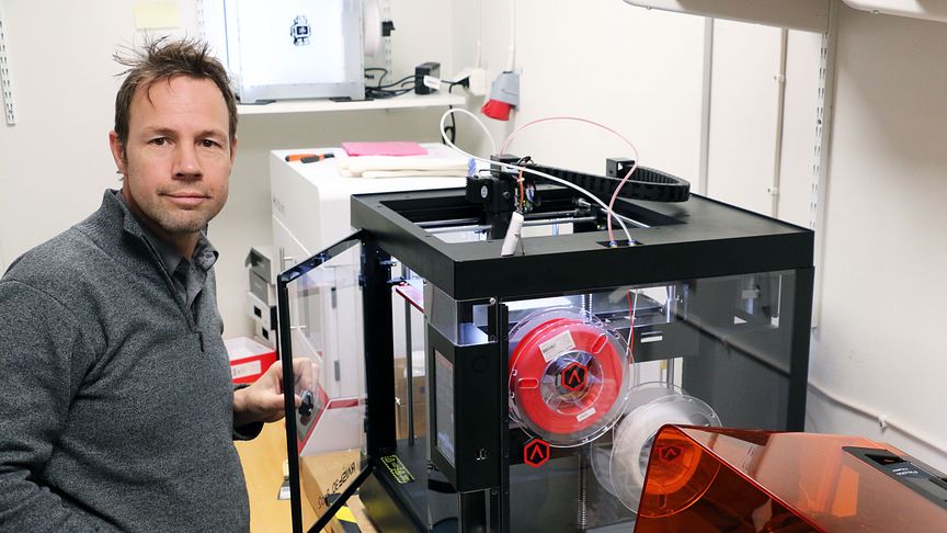 Einar Heiberg ses till vänster i bilden. Han är klädd i grå tröja och har brunt kortklippt hår. Han öppnar luckan till en fyrkantig box med genomskinliga sidor. På sidan är fästade två rullar av vad som verkar vara slangar. Den ena rullen är röd och den andra vit. Boxen föreställer 3D-printern