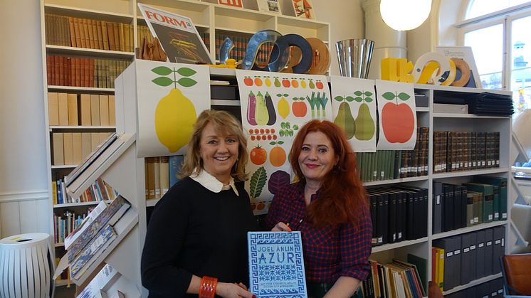 ​Ewa Kumlin och Lotta Kühlhorn i biblioteket på Svensk Form, med vinnarboken AZUR framför de prisvinnande affischer som Lotta har gjort för Akademibokhandeln.