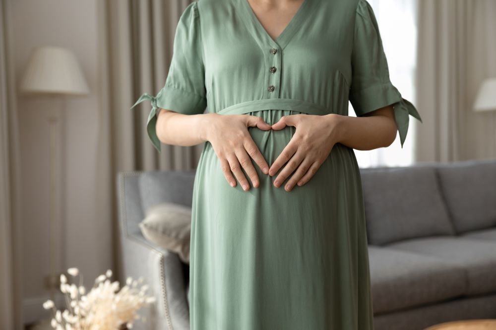 Syntolkning: Närbild på gravid kvinnas mage och hennes händer på magen i hemmiljö.