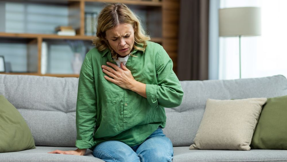 Syntolkning: Kvinna som har ont i bröstkorgen sitter på soffa med handen på bröstet